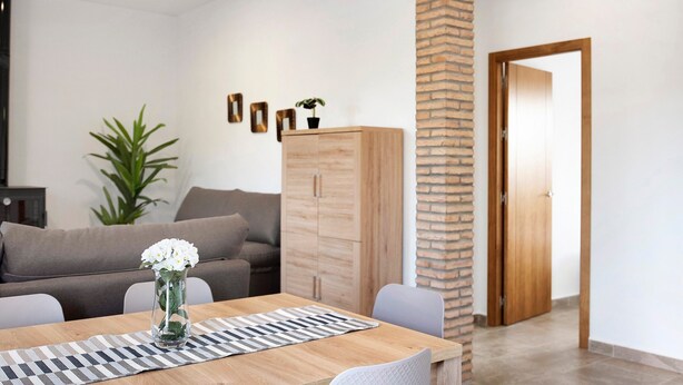 A living room with a sofa, a wooden cabinet, a dining table with a placemat, and a potted plant. - Holiday Home "Villa Montenmedio - Paula" with Mountain View & Pool (El Soto)