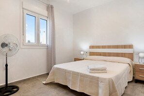 A bedroom with a bed, bedside table, lamp, and a standing fan.