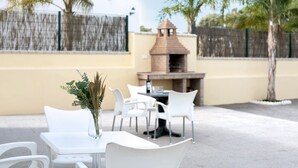 An outdoor patio with white chairs and a table, a brick barbecue, and a decorative fence.