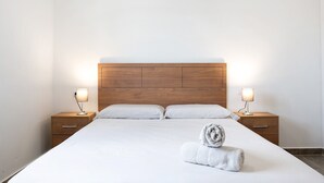 A neatly made bed with two bedside tables, each with a lamp, in a room with a wooden headboard and plain walls.