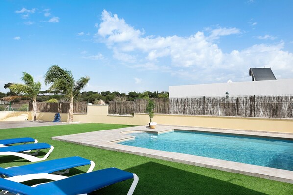 A swimming pool with blue lounge chairs, a small palm tree, and a white building in the background.