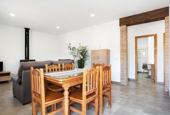A dining area with a wooden table and chairs, a sofa, a potted plant, and a brick column.