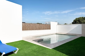 A swimming pool with a blue lounge chair, surrounded by artificial grass and a white wall.