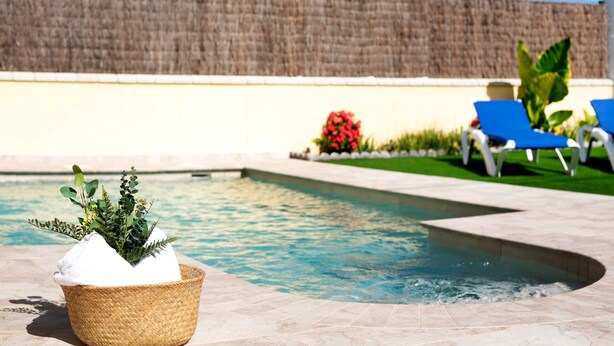 Une piscine, avec un panier en osier contenant une plante et une serviette, entourée de chaises longues bleues et d’un mur en pierre.
