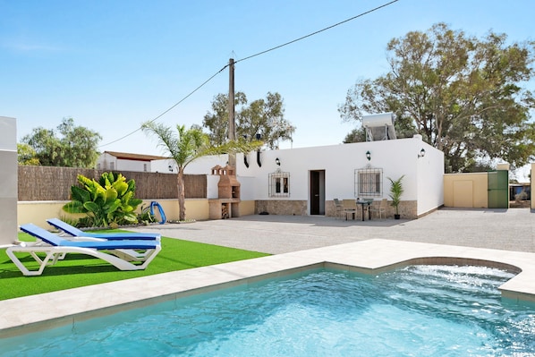 Une piscine avec des chaises longues, un espace patio et un bâtiment blanc doté d’une cheminée.
