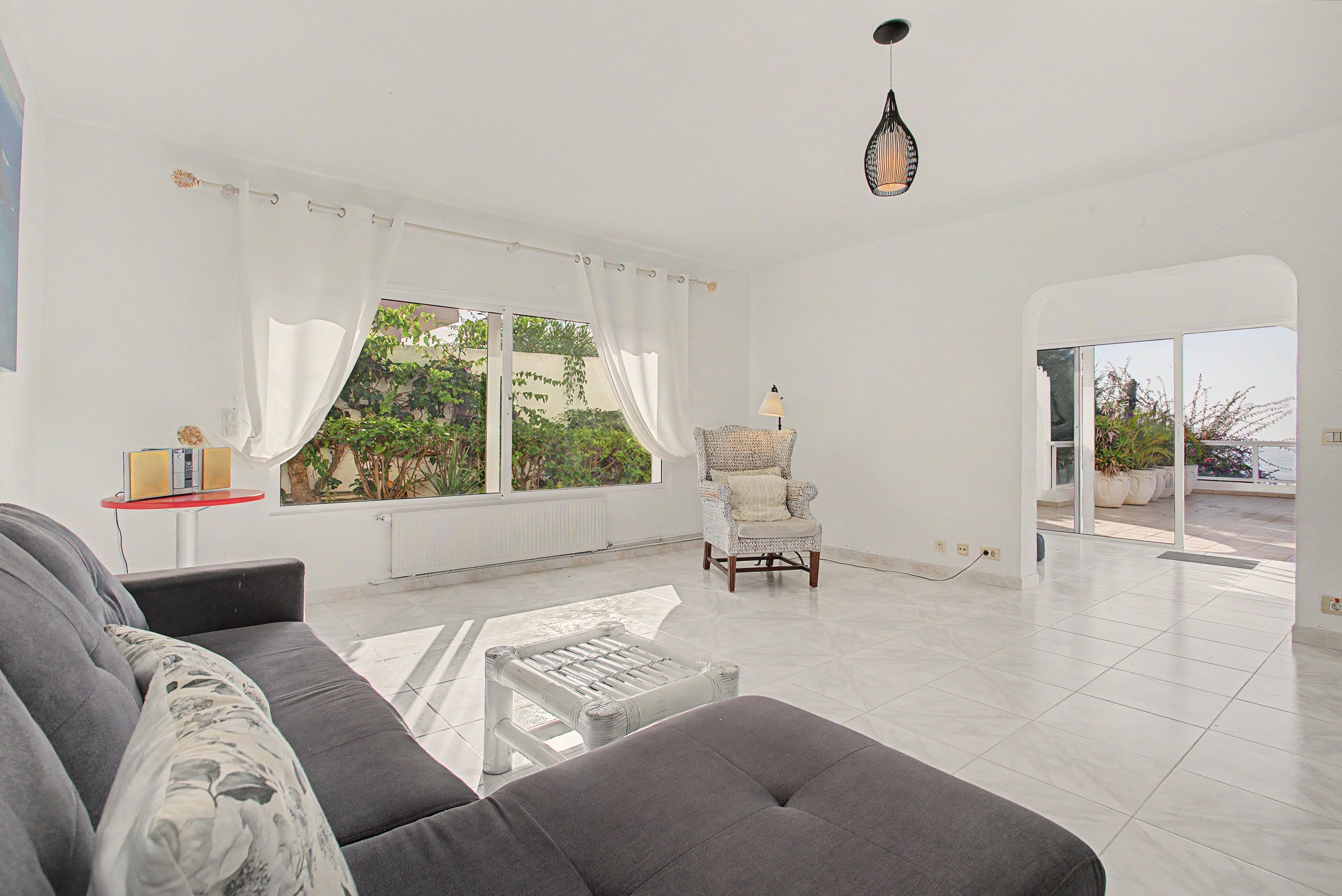 A spacious living room with a sofa, a chair, a table, and a view of greenery through large windows.