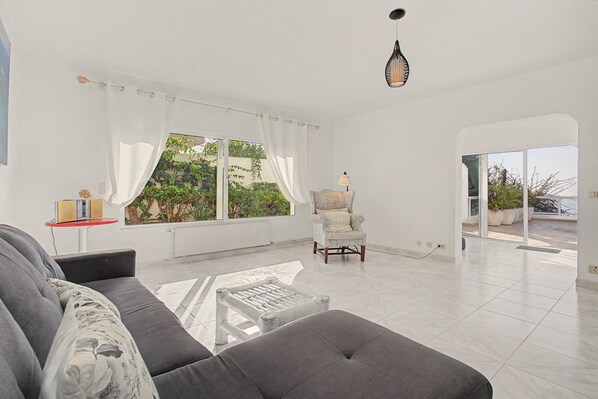 A spacious living room with a sofa, a chair, a table, and a view of greenery through large windows.