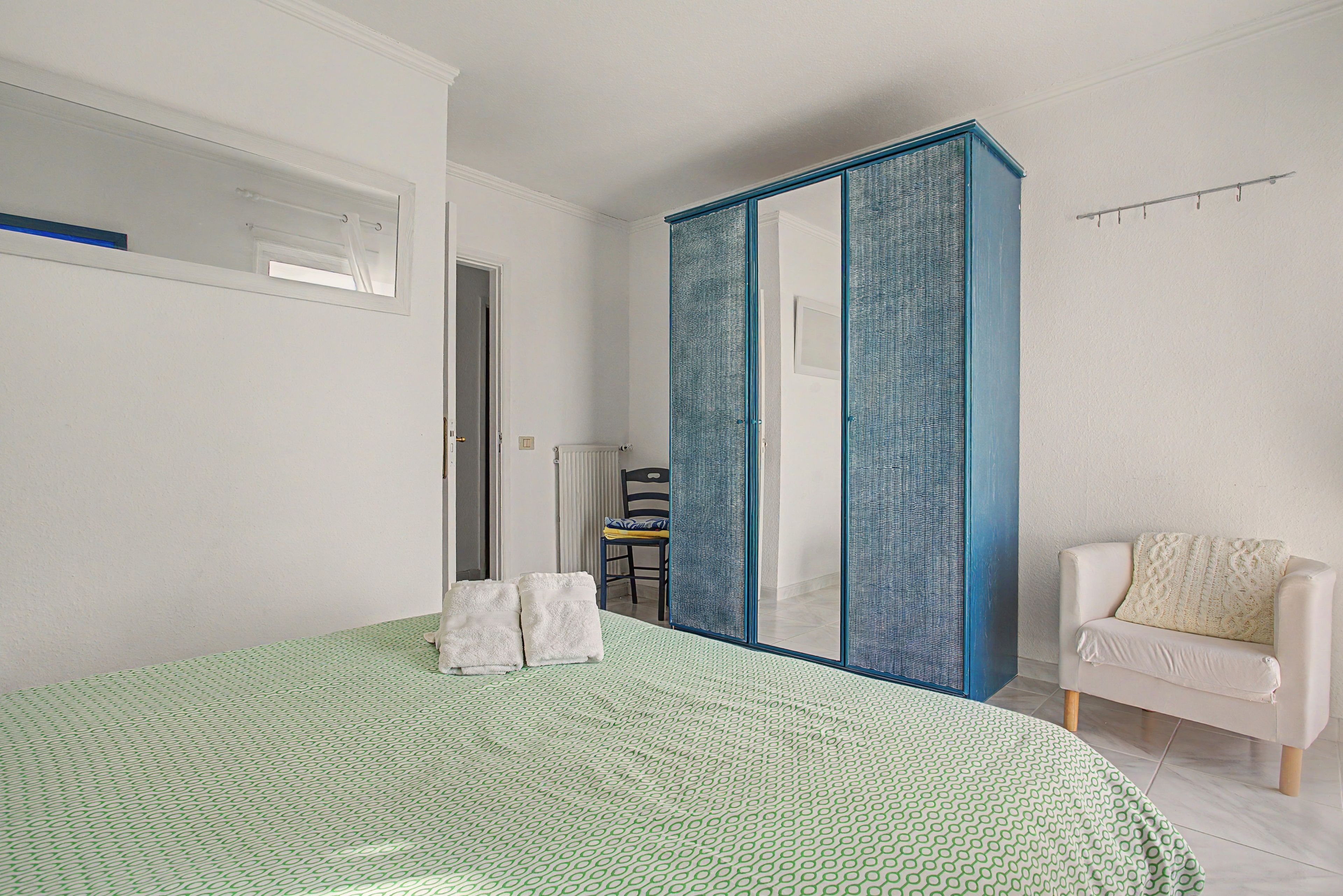 A bedroom with a bed, a white chair, a blue wardrobe, and a green patterned rug.