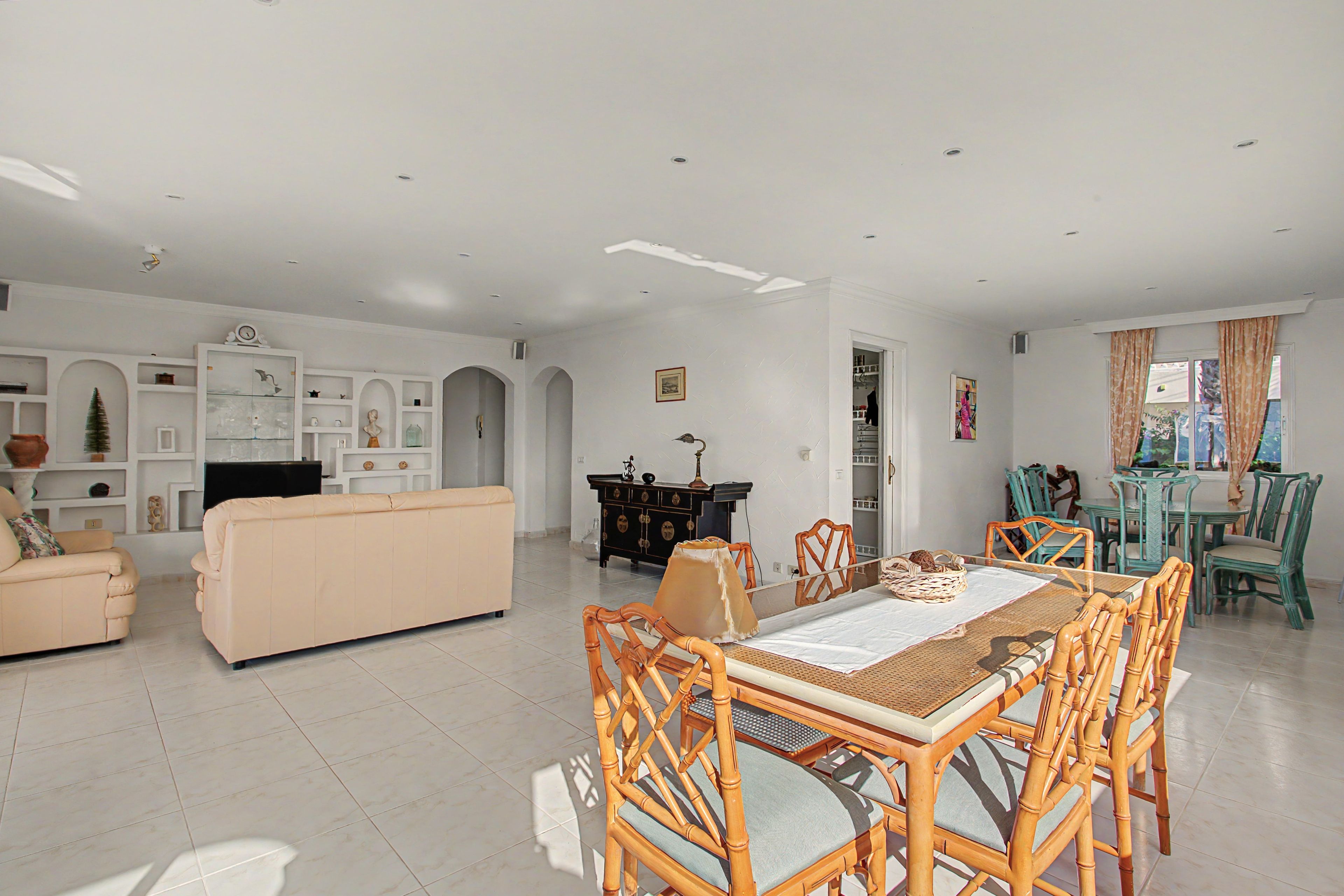 A spacious living room with a beige sofa, a dining table with bamboo chairs, and a kitchen area in the background.