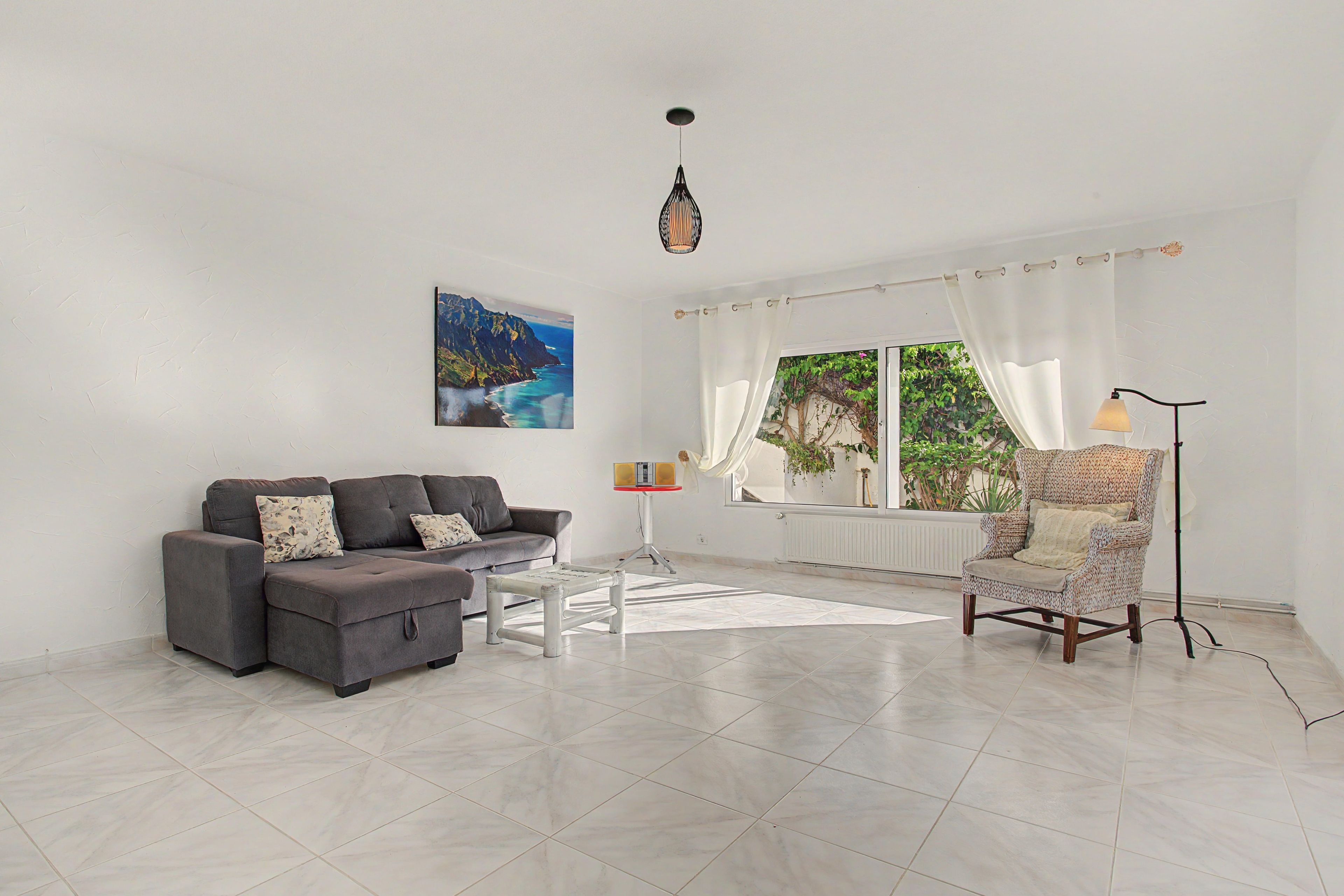 A spacious living room with a grey sofa, a patterned armchair, a coffee table, and a large window with curtains.