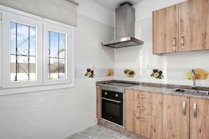 Eine Küche mit weißen Fliesen, einer Edelstahl-Dunstabzugshaube, Holzschränken, einem eingebauten Backofen und einem Fenster mit schmiedeeisernen Gitterstäben.