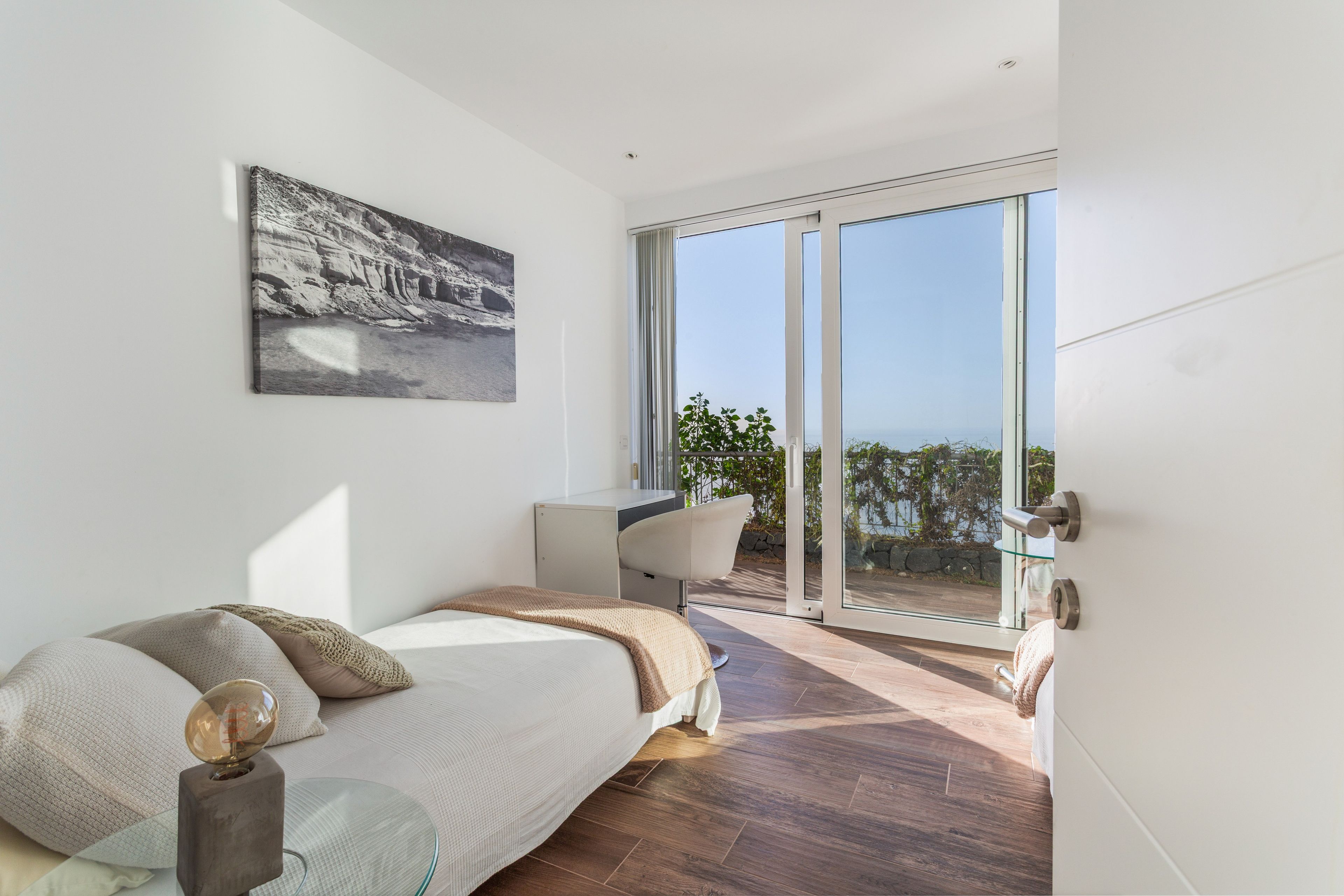 A modern bedroom with a bed, a desk, and a large sliding glass door leading to a balcony with a view.