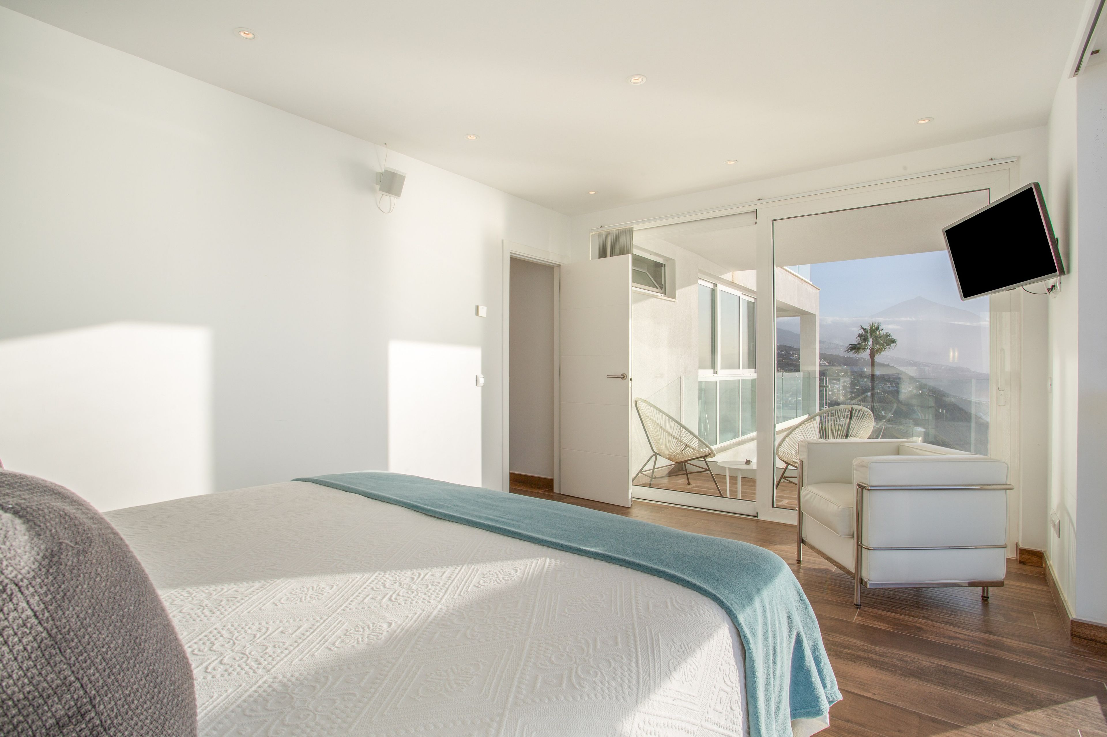 Quarto moderno com cama grande, área de estar com poltrona e mesa, TV fixada na parede e varanda com vista.
