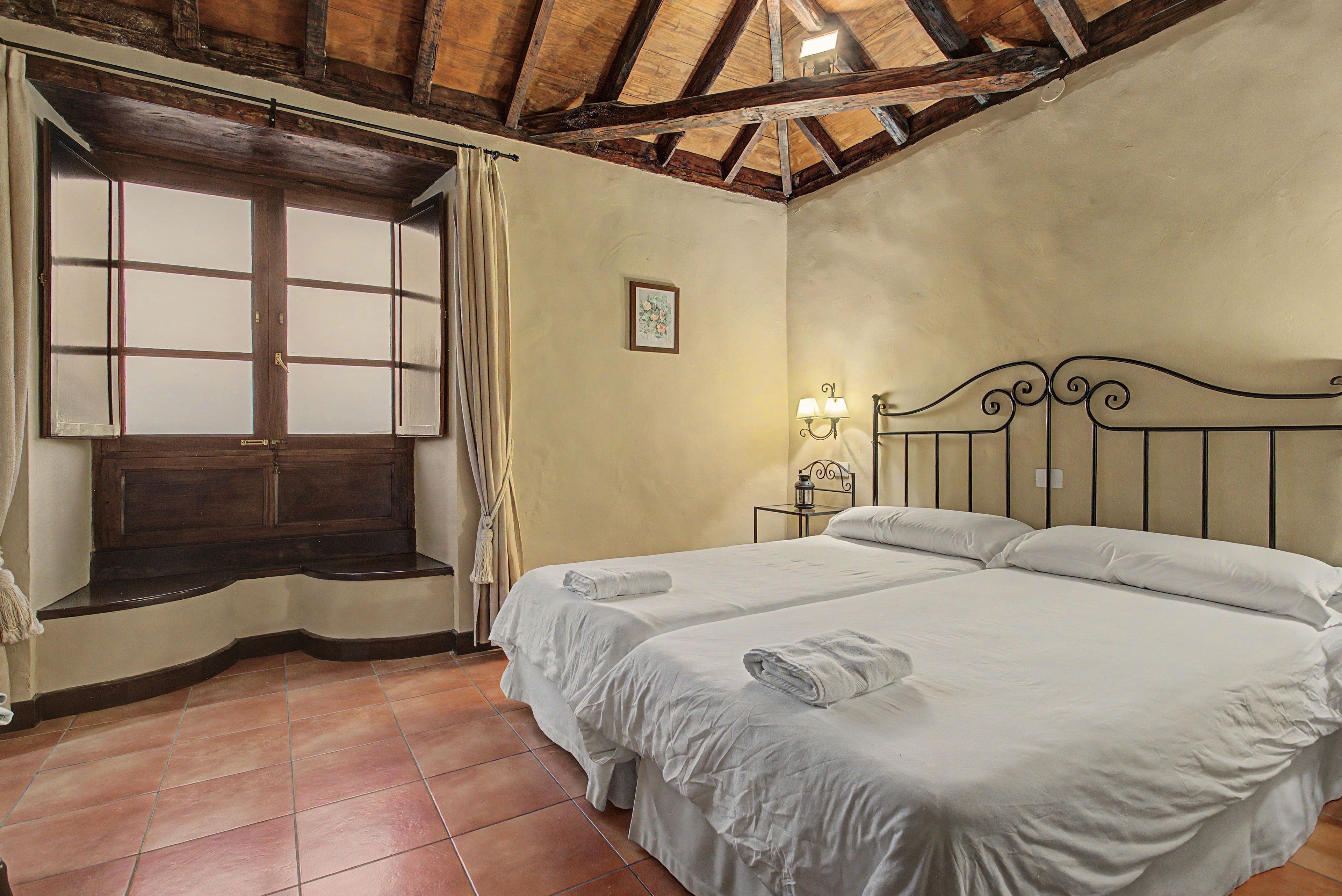 Quarto com duas camas, janela com cortinas, mesa de cabeceira e abajur, e teto de madeira.