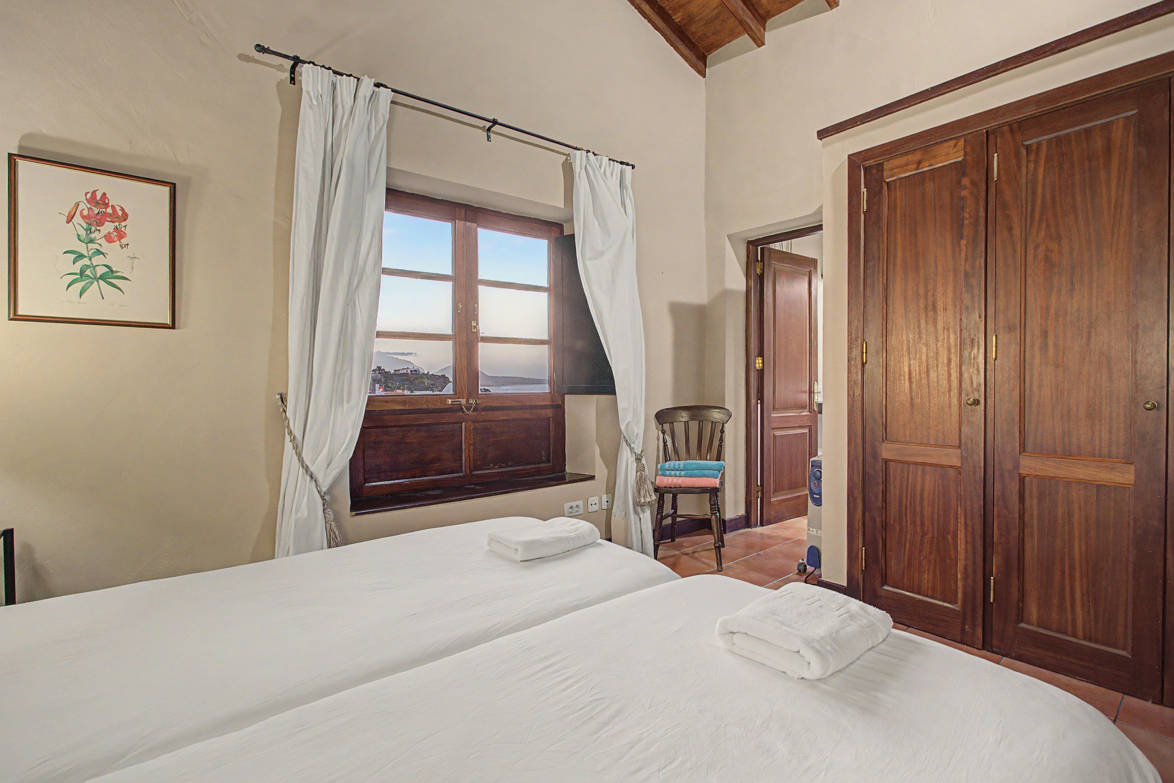 Ein Schlafzimmer mit einem Bett, einem Fenster mit Vorhängen, einer Holztür und einem gerahmten Bild an der Wand.