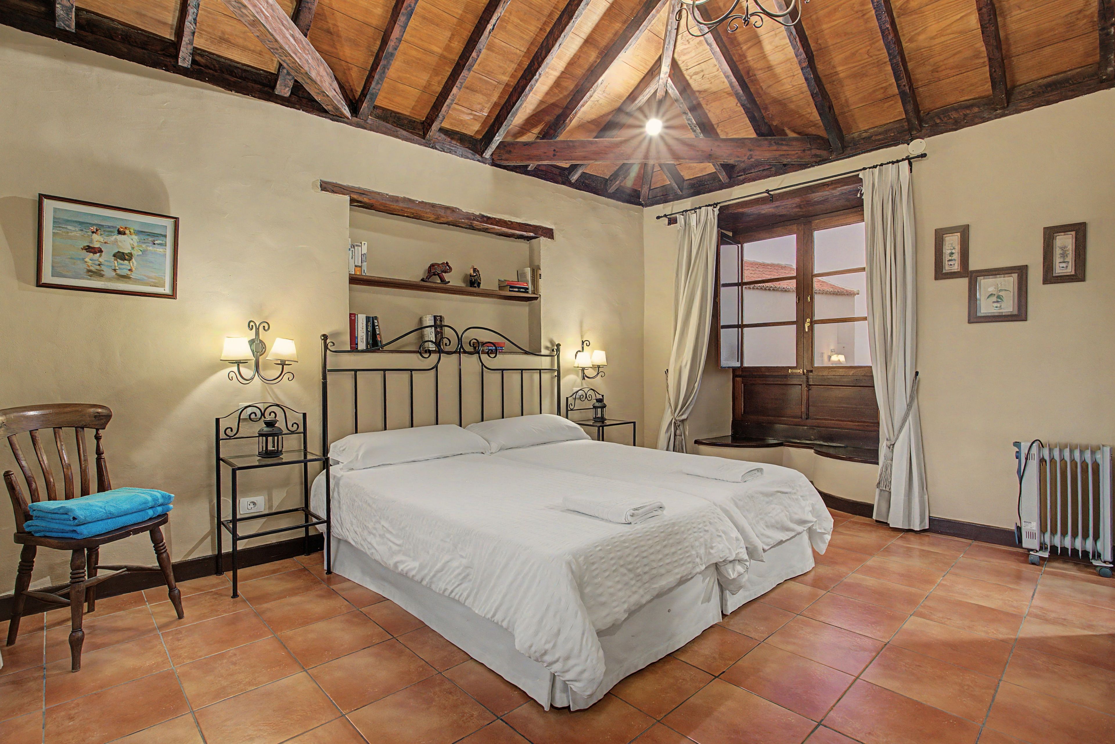Un dormitori amb un llit gran, una cadira, una petita taula de nit, una estantària amb objectes decoratius, una finestra amb cortines i un radiador.