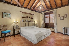 Een slaapkamer met een groot bed, een stoel, een nachtkastje, een plank met decoratieve spullen, een raam met gordijnen en een radiator.