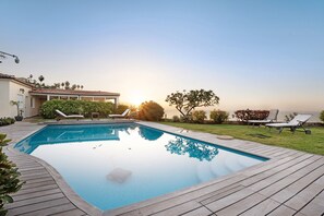A swimming pool with lounge chairs, a pergola, and a view of the sunset.