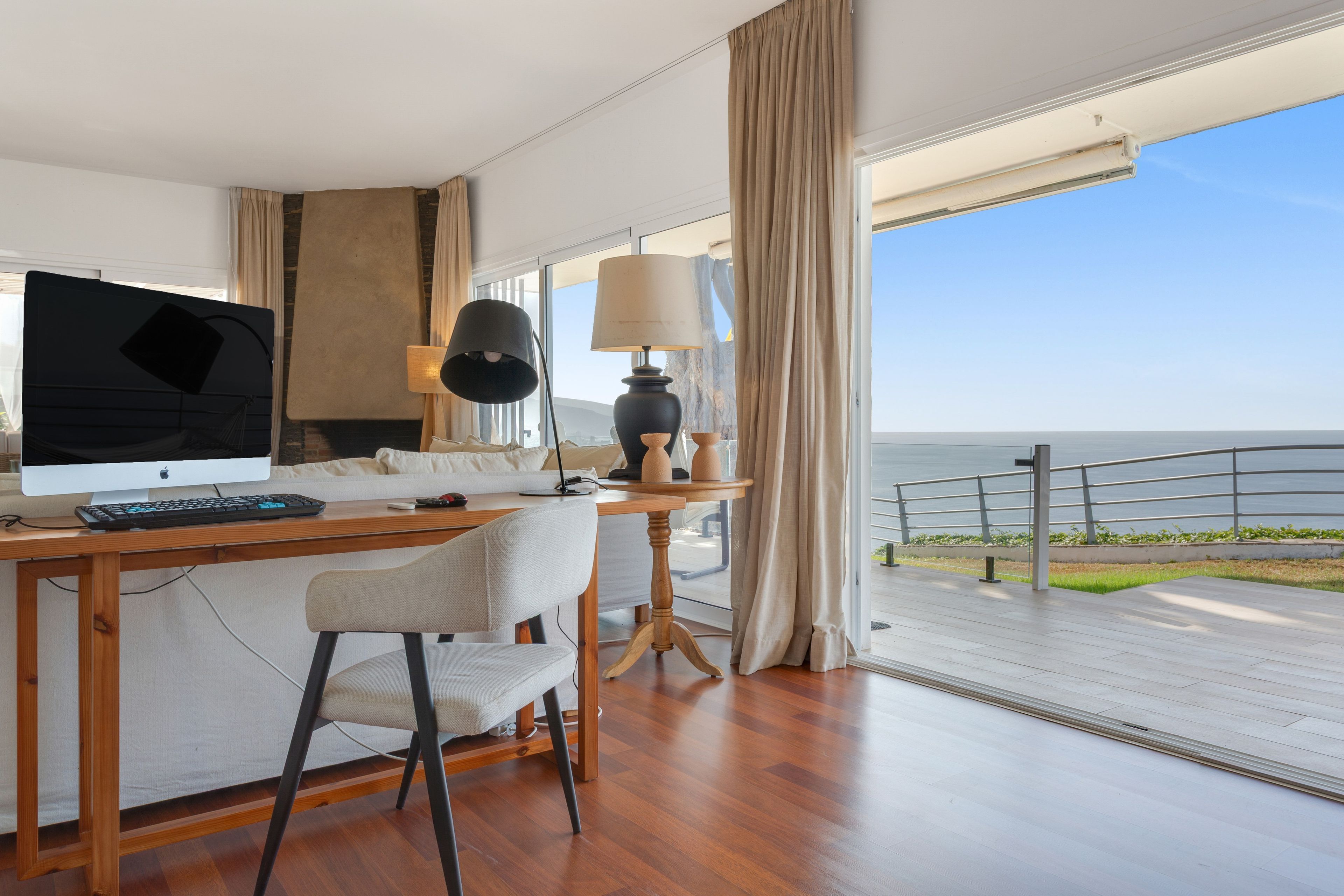 Een moderne kamer met een houten bureau, een computer, een stoel, een lamp en uitzicht op zee.