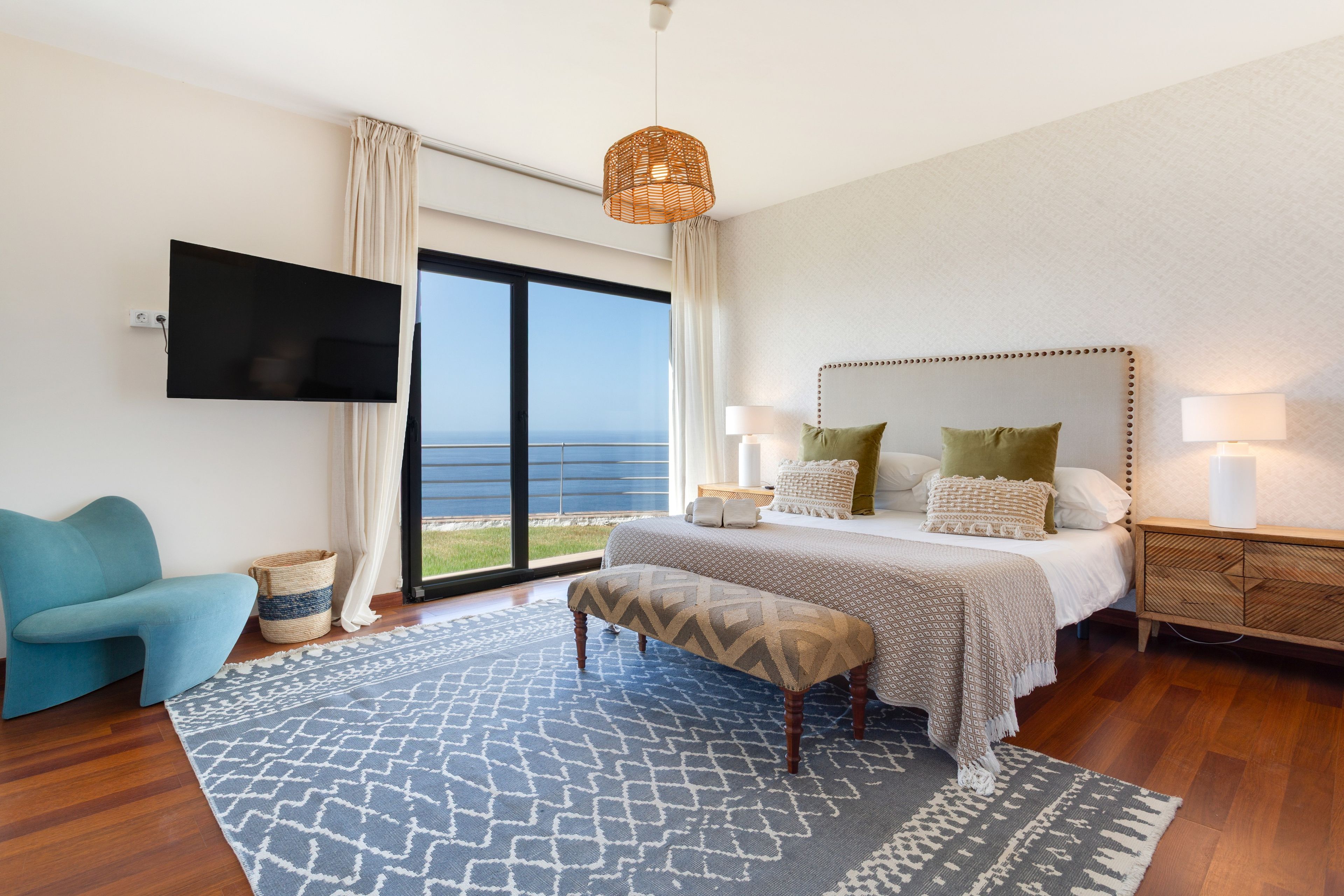 A bedroom with a large bed, a television mounted on the wall, and a view of the sea through the window.