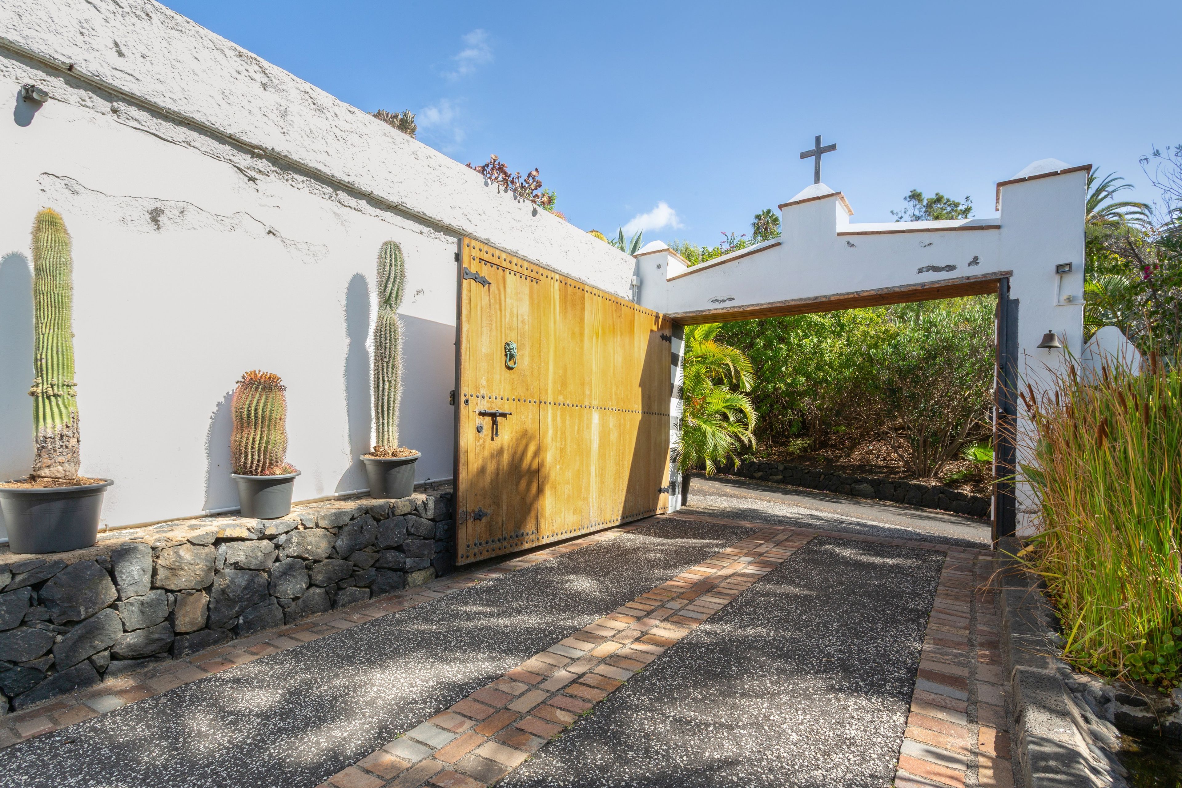 A gated entrance with a wooden door, a white wall with cacti, and a stone pathway leading to a building with a cross on top.