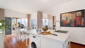 A modern kitchen with a built-in washing machine, a dining area with white chairs, and a large window with curtains. - Vacation Home "Villa Bahía de Michel" with Sea View, Pool, Garden & Wi-Fi (santa ursula)
