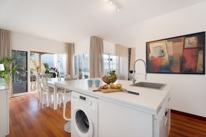 Une cuisine moderne dotée d’un lave-linge encastré, d’un coin repas avec des chaises blanches et d’une grande fenêtre agrémentée de rideaux.