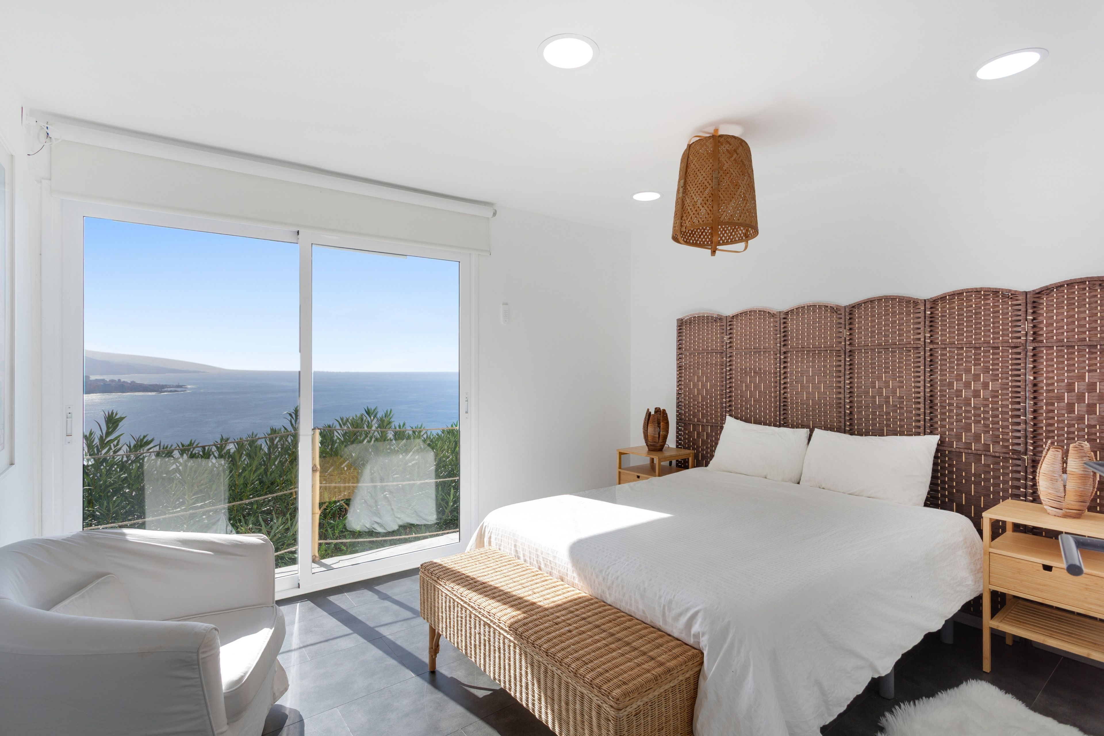 A bedroom with a large bed, a nightstand, a wicker bench, and a view of the sea.
