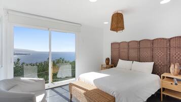 A bedroom with a large bed, a nightstand, a wicker bench, and a view of the sea.