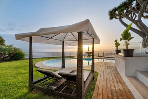 Un espace aménagé au bord de la piscine, comprenant un coin salon couvert, une terrasse en bois et une vue sur l’océan.