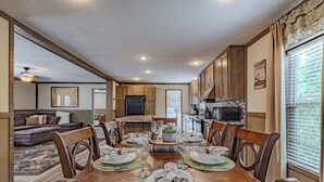 Un comedor amplio con una mesa de madera dispuesta para seis personas, rodeada de sillas de madera. Se ve una cocina con armarios de madera y un microondas al fondo.