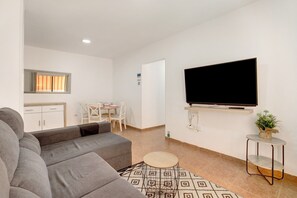 Un salon moderne avec un canapé gris, un téléviseur fixé au mur, une petite table ronde et, au fond, un coin repas.