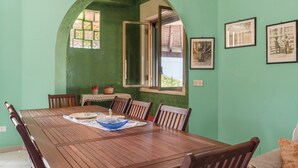 Een eethoek met een houten tafel, stoelen, een raam met open luik en ingelijste foto's aan de muur.