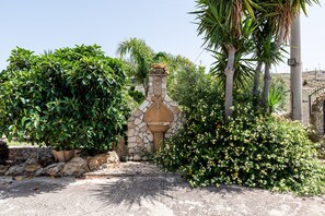 Una fuente de piedra con una parte superior decorativa, rodeada de abundante follaje y plantas florecientes.