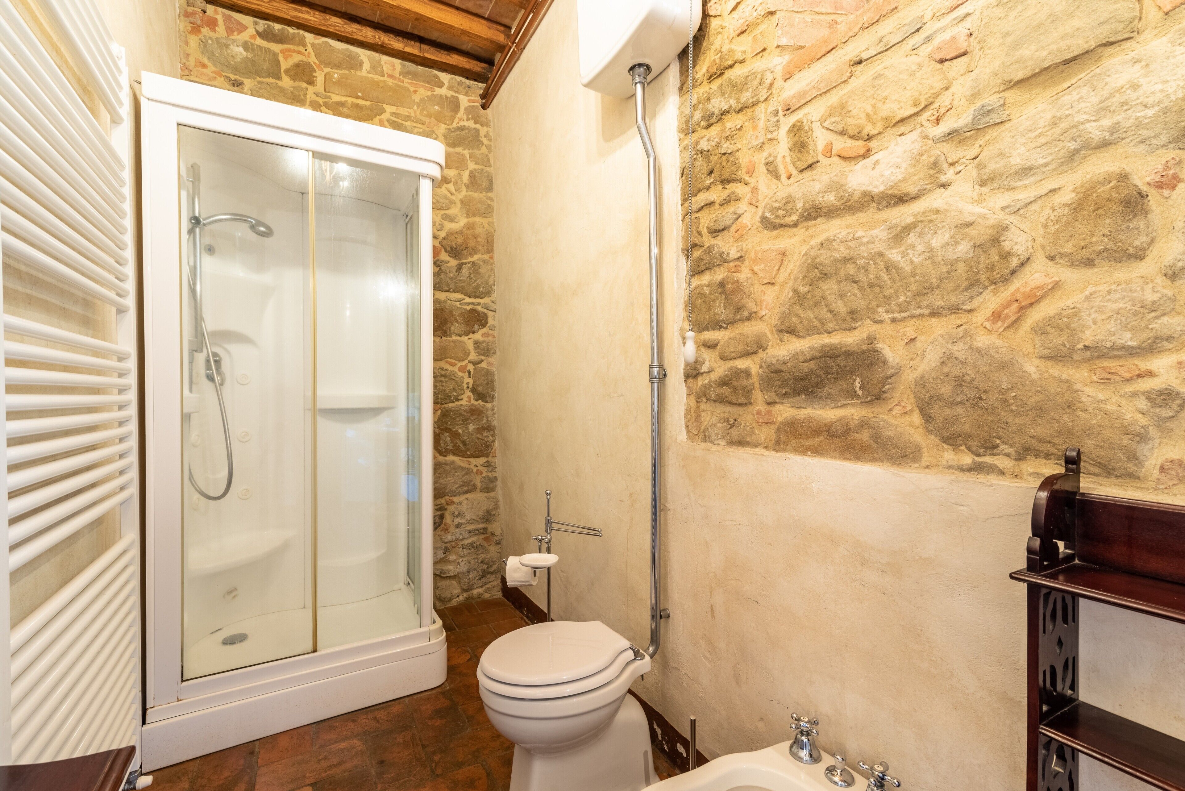 A bathroom with a shower, toilet, and sink, featuring stone walls and wooden beams.