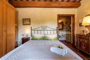 A bedroom with a bed, wooden wardrobe, bedside table, lamp, and a stone wall.