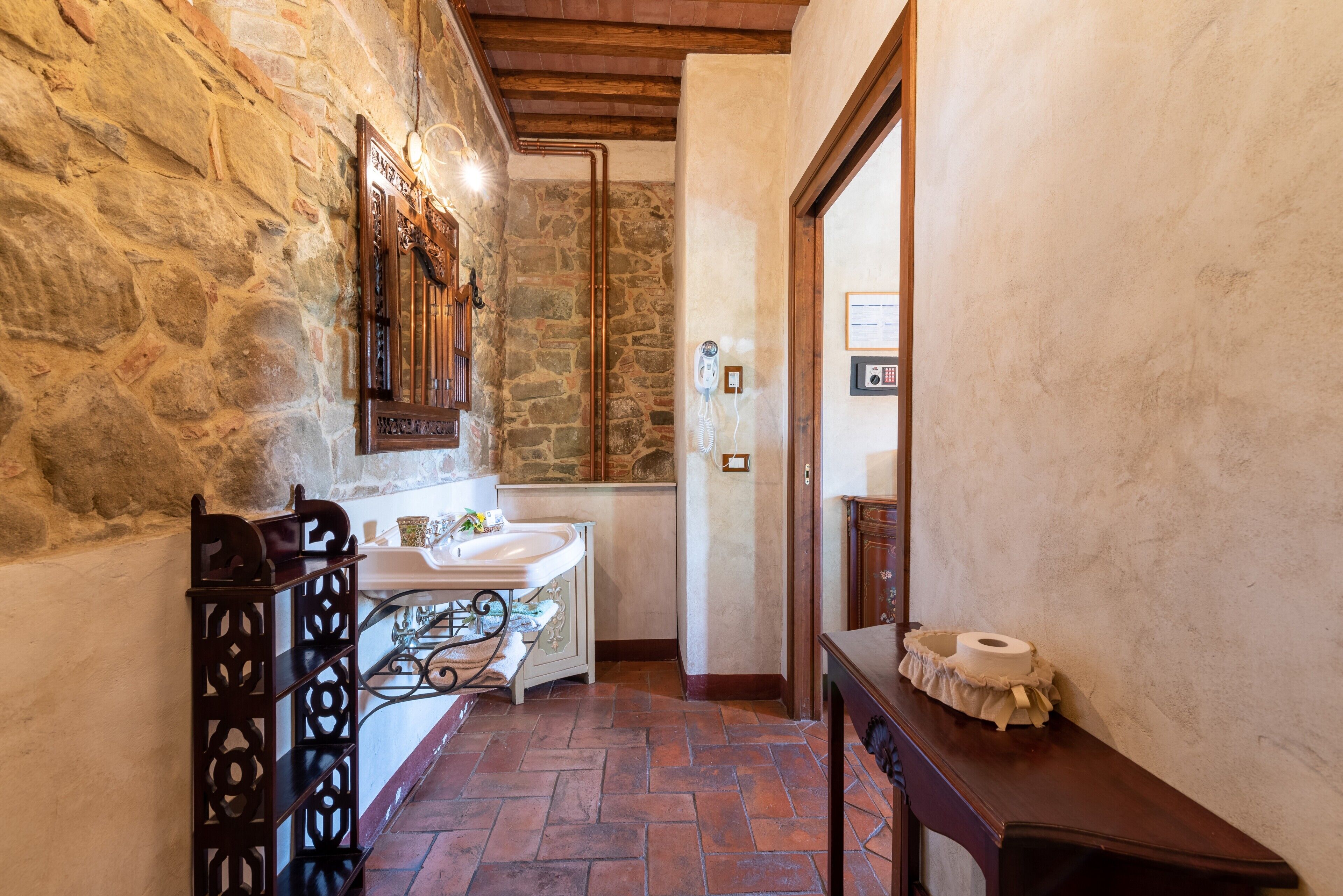 A bathroom with a stone wall, wooden beams, a sink, a mirror, and a toilet paper holder.