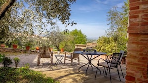Un patio exterior con mesa y sillas, rodeado de olivos y un muro de piedra.