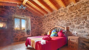 A bedroom with a stone wall, wooden ceiling beams, a bed with colorful pillows, a nightstand, and a dresser.