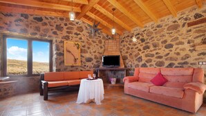A stone-walled room with a wooden ceiling, a stone fireplace, a television, a pink sofa, a white table with glasses, and a bench.