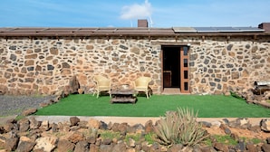 A stone building with a green lawn, two wicker chairs, a wooden table, and a barrel.