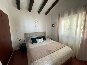Une chambre avec un lit, une table de chevet, un attrape-rêves fixé au mur, une fenêtre avec des rideaux et des poutres en bois apparentes au plafond.