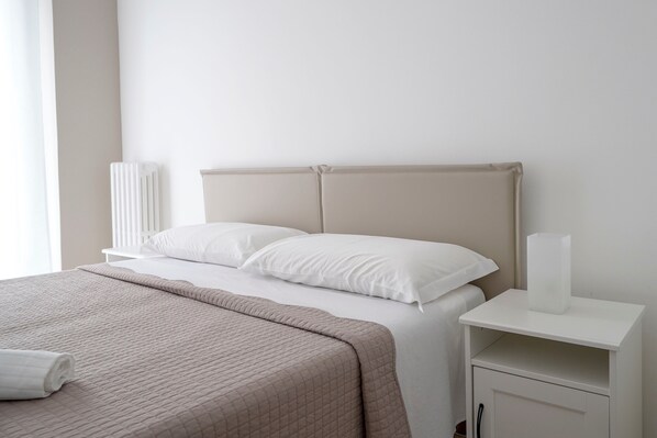 A neatly made bed with white bedding, a beige headboard, and a white bedside table with a lamp.