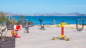 Uma área de exercícios ao ar livre com diversos equipamentos de ginástica, vista desobstruída para o mar e montanhas ao fundo.