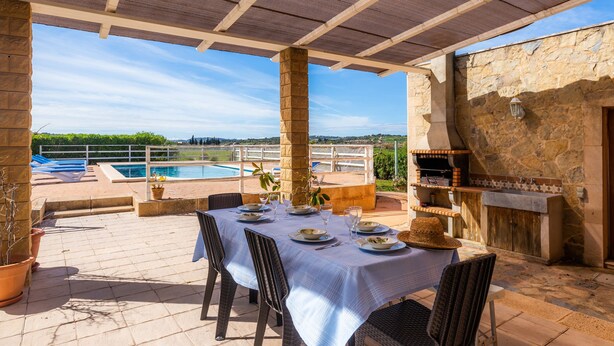 An outdoor patio with a table set for dining, a stone wall, and a pool in the background. - Casa "Cova Dor" with Shared Pool, Garden and Wi-Fi (Santa Margalida)