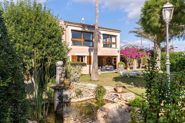 Una casa de dos pisos con jardín, que incluye un estanque, muebles de exterior y un farol.