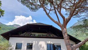 A white building with a sloped roof and wooden beams, surrounded by greenery and mountains.