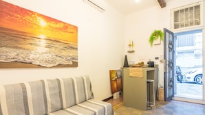 A room with a sofa, a painting of a sunset, a small desk with a plant, and a view of the street outside. - Apartment 'Casa Pepes Centro Ortigia' with Shared Garden, Wi-Fi and Air Conditioning (Siracusa)