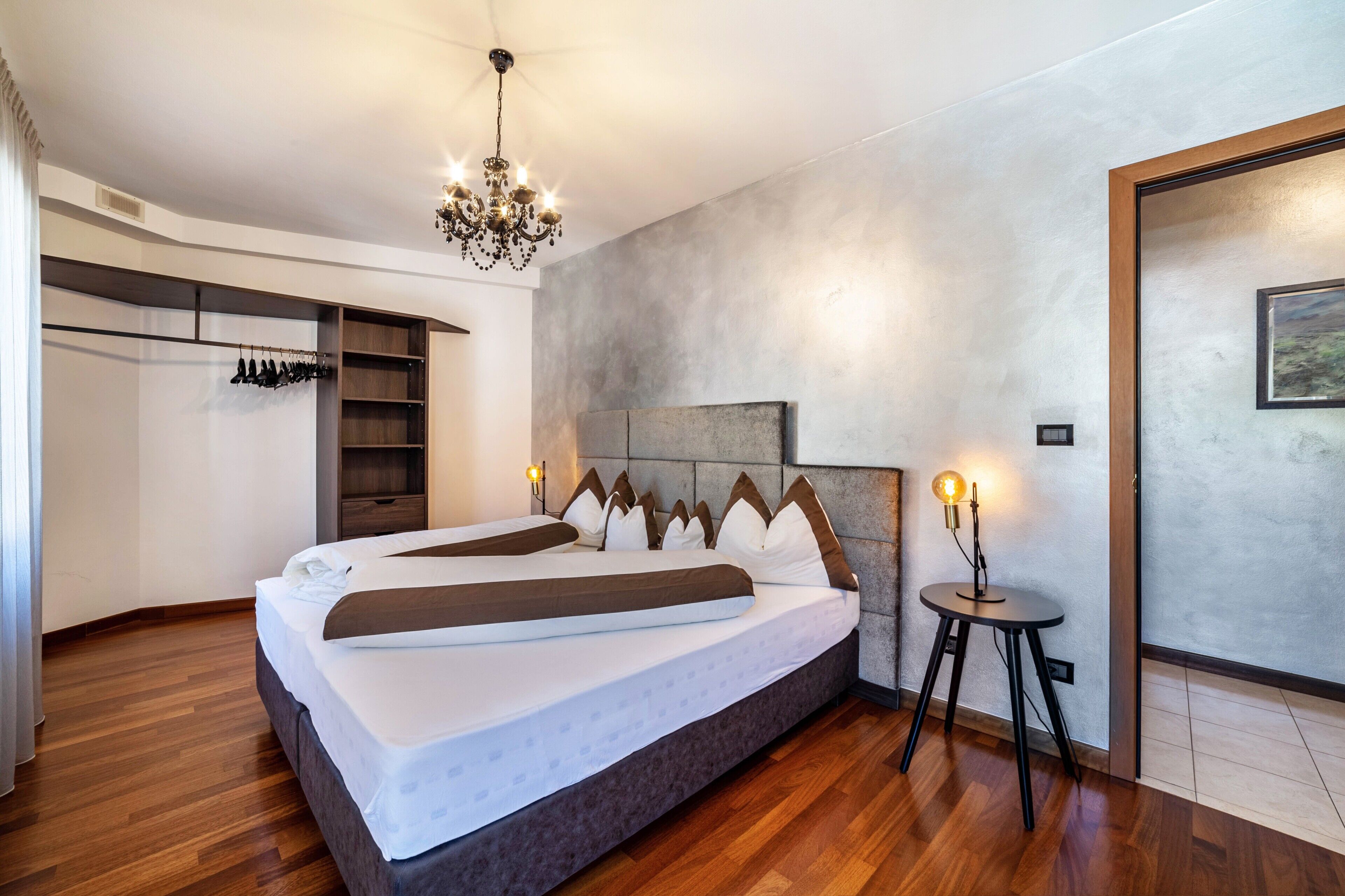 Une chambre à coucher moderne avec un grand lit, un lustre, une petite table avec une lampe et une armoire où sont suspendus des vêtements.