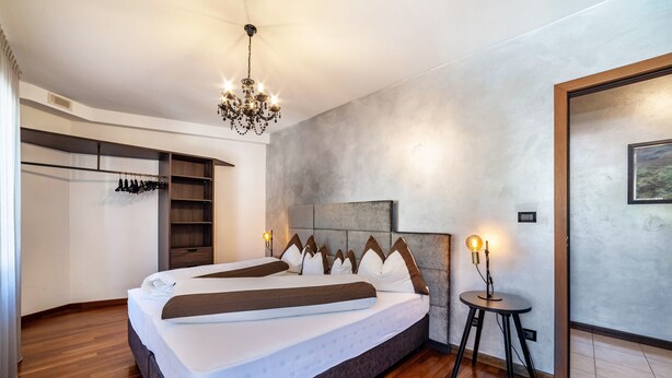 A modern bedroom with a large bed, a chandelier, a small table with a lamp, and a wardrobe with hanging items. - Holiday Apartment 'König - The King 4th' with Balcony & Wi-Fi (Meran)
